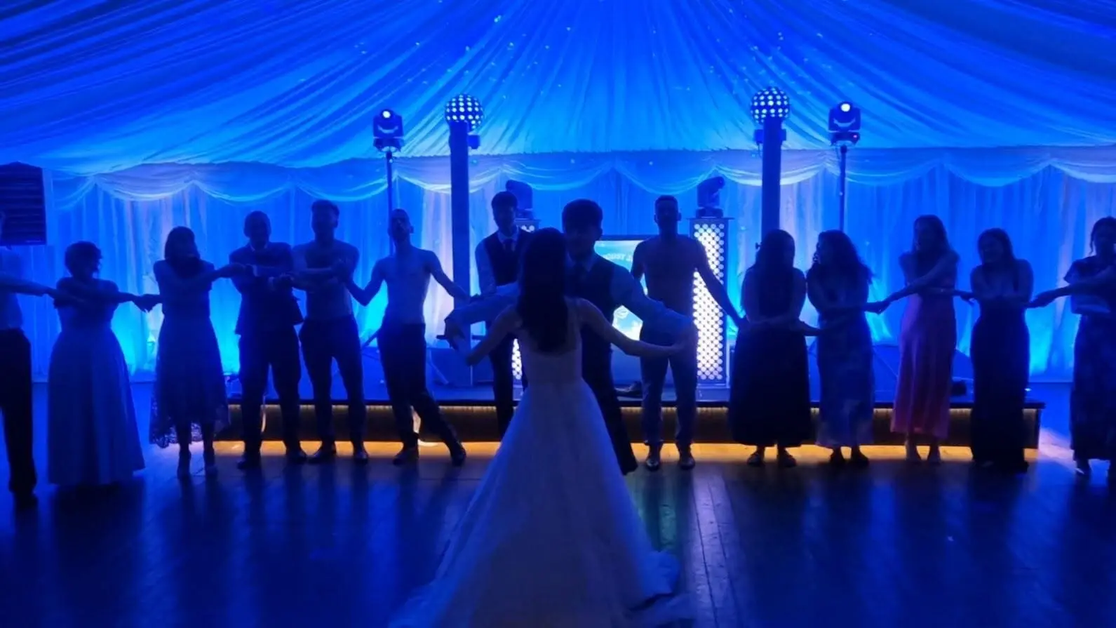 A bride and groom dancing in front of a line of their guests holding hands, lit blue in a marquee