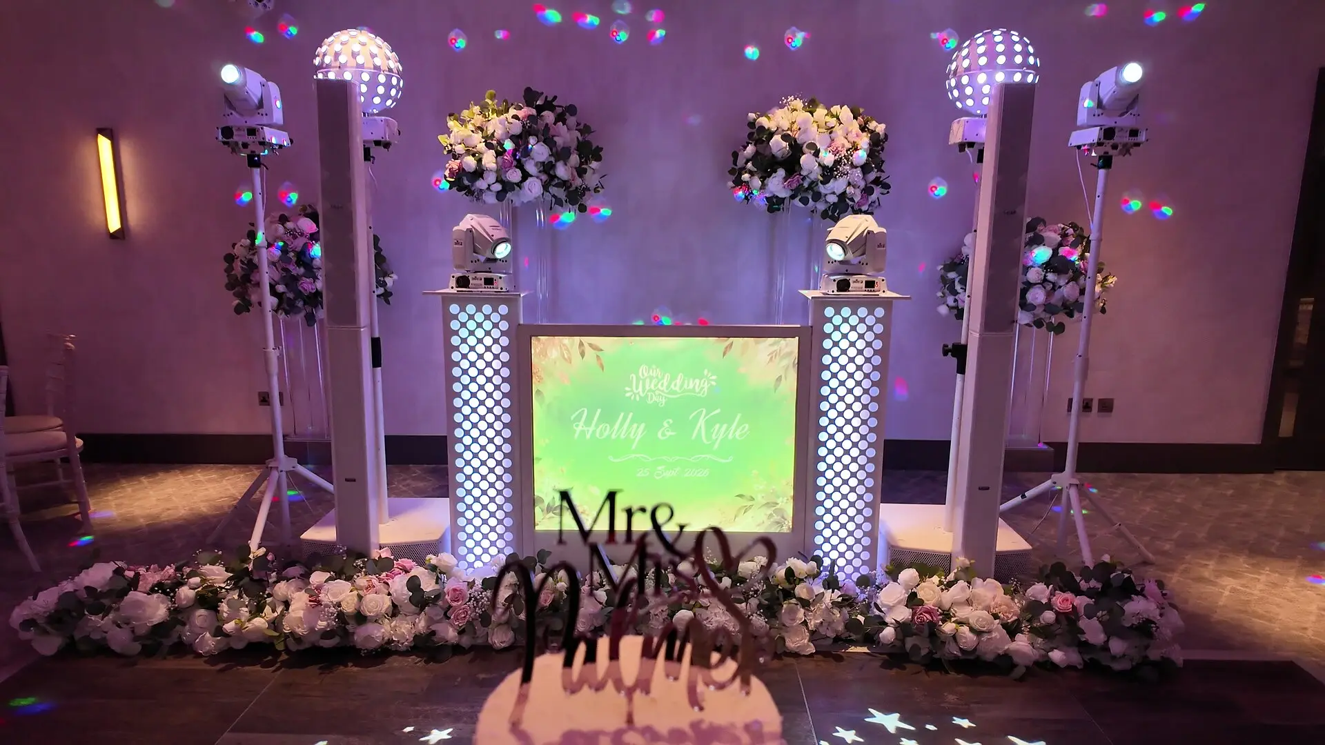 The all-white DJ setup framed by flowers, its screen showing the couple's names in a bespoke design